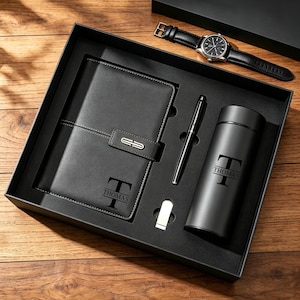 Könnte beinhalten: Ein schwarzes Geschenkset in einer Box mit einem lederartigen Notizbuch mit dem Namen THOMAS und einem T-Monogramm, einem schwarzen Stift, einem kleinen silbernen Clip, einer schwarzen zylindrischen Flasche mit dem Namen THOMAS und einem T-Monogramm sowie einer schwarzen Ledernuhr.