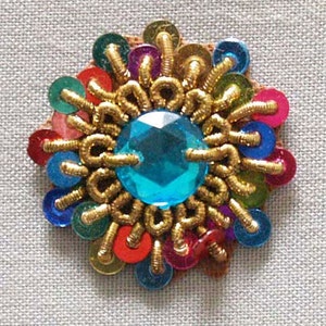 May include: A colorful, decorative embellishment with a central, faceted, teal-colored gem. The design features gold-colored thread and a ring of multi-colored sequins in red, blue, green, pink, and purple. The embellishment is likely for clothing or crafts.