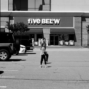 May include: A black and white photograph of a Five Below retail store exterior with a parking lot in the foreground. Two individuals are walking across the pavement toward the store entrance, which features signage displaying the text Five Below.