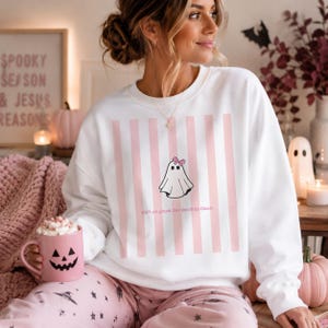 Può includere: Una felpa bianca a girocollo con una grafica centrale raffigurante un fantasma con un piccolo fiocco rosa, su uno sfondo a righe verticali rosa chiaro. Il testo "ain't no ghost like the Holy Ghost" è stampato in piccole lettere rosa sotto il fantasma.