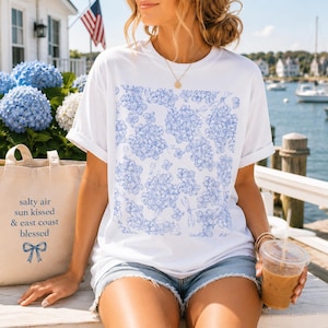 May include: A white short-sleeve t-shirt featuring a blue floral pattern on the front, paired with a beige canvas tote bag printed with the text "salty air sun kissed & east coast blessed" and a blue bow icon, alongside a plastic cup of iced coffee.
