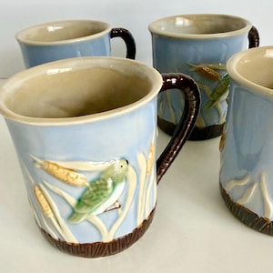 May include: A set of four light blue ceramic mugs featuring a raised relief design of green birds perched among tan cattails and reeds. Each mug has a dark brown textured base and a matching dark brown handle with a smooth, glossy finish.