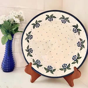 May include: A round ceramic plate featuring a cream-colored base with a pattern of small dark blue dots and a border of blue rose motifs with green leaves. The plate has a dark blue rim and is displayed on a wooden stand next to a blue vase.