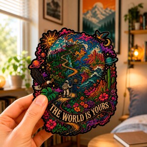 Puede incluir: Una pegatina troquelada con una vibrante ilustración psicodélica de la Tierra con un camino sinuoso, un astronauta sosteniendo una bandera, un águila, un sol y planetas. El texto THE WORLD IS YOURS está impreso en una cinta en la parte inferior.