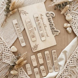 Peut inclure: Une plaque en bois clair portant le prénom "Swaeli" et le texte "Aujourd'hui c'est mon premier jour en petite section" et "mon dernier jour", accompagnée de plusieurs petites étiquettes en bois indiquant les niveaux scolaires français.