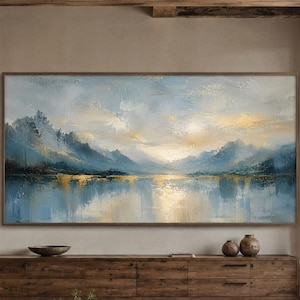 May include: A large rectangular landscape painting featuring a serene mountain range and a calm lake with soft blue, gold, and white brushstrokes. The artwork is displayed in a wooden frame and mounted on a neutral wall above a long wooden console table.