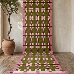 Könnte beinhalten: Ein langer, rechteckiger Läufer mit einem sich wiederholenden geometrischen Muster in Olivgrün, Creme und Dunkelrot. Der Teppich ist von einem breiten rosa Streifen umrandet und schließt mit einer dunkelroten Borte und beigen Naturfaserfransen ab.