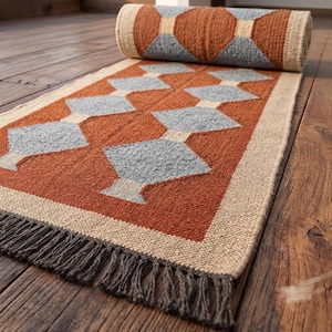 May include: A rectangular woven runner rug featuring a geometric pattern of light blue diamonds on a burnt orange background. The rug has a thick beige border and dark brown fringe at the end. A portion of the rug is rolled up at the top edge.