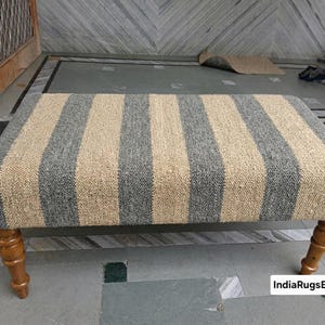Op de afbeelding: Een rechthoekige bank met een geweven jute bovenkant met afwisselende verticale strepen in natuurlijk beige en donkergrijs. De bank wordt ondersteund door vier gedraaide houten poten met een warme bruine afwerking. De tekst IndiaRugsExport is zichtbaar.