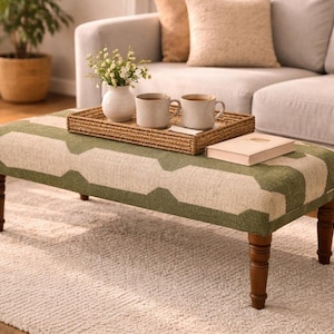 May include: A rectangular ottoman featuring a green and cream geometric pattern, supported by four dark brown turned wooden legs. A woven rectangular tray sits on top, holding a white vase with small flowers and two light-coloured ceramic mugs.