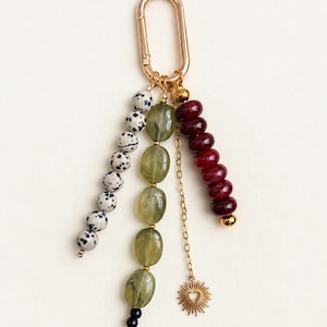 May include: A gold-toned oval carabiner keychain featuring three hanging strands. One strand has white and black spotted beads, another has translucent olive green oval beads with a small black bead, and the third has deep red beads and a sun charm.