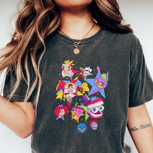 May include: A charcoal gray short-sleeve t-shirt featuring a colorful graphic print of various cartoon characters, including a jester, a star, and a toothy creature, arranged in a cluster on the front center of the garment.