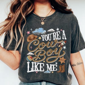 May include: A charcoal gray short-sleeve t-shirt featuring a graphic print with the text "YOU'RE A COWBOY LIKE ME" in white and rope-textured lettering. The design includes a brown hat, a rocket, a sheriff badge, boots, and small stars.