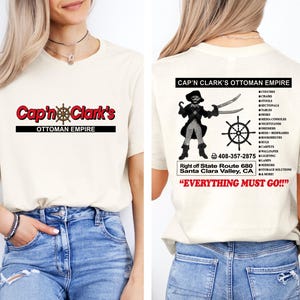 May include: A cream-colored t-shirt featuring a front graphic with the red text Cap'n Clark's and a ship wheel. The back displays a pirate illustration, the text Cap'n Clark's Ottoman Empire, a list of furniture, a phone number, and the text EVERYTHING MUST GO!!