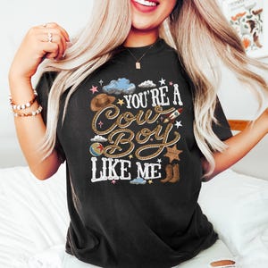 May include: A black t-shirt featuring a graphic print with the text "YOU'RE A COW BOY LIKE ME" in a rope-style font. The design includes illustrations of a brown cowboy hat, a rocket, a ball, a sheriff star badge, brown boots, and blue clouds.