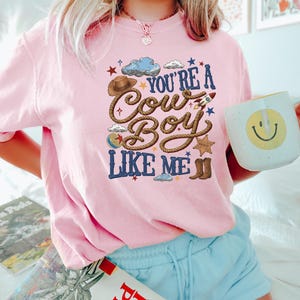 May include: A light pink t-shirt featuring a graphic print with the text "YOU'RE A COWBOY LIKE ME" in blue letters. The design includes a brown cowboy hat, a rope, a small rocket, a sheriff star badge, brown boots, a ball, and blue clouds.