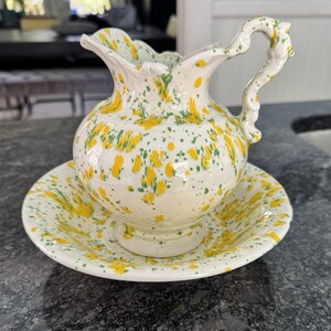 Könnte beinhalten: Ein Keramik-Krug mit passender Untertasse, bestehend aus einer cremefarbenen Basis mit einem gesprenkelten Muster aus leuchtend gelben und grünen Farbspritzern. Der Krug hat einen gewellten Rand, einen Standfuß und einen verzierten Griff.