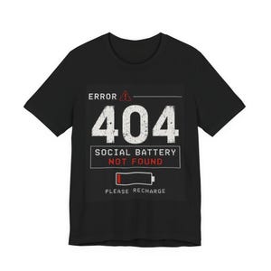 以下が含まれることがあります： ERROR 404、SOCIAL BATTERY NOT FOUND、PLEASE RECHARGEというテキストがプリントされた黒の半袖Tシャツ。デザインには、赤い警告三角形のアイコンと、赤いバーが表示されたバッテリーのシンボルが含まれています。