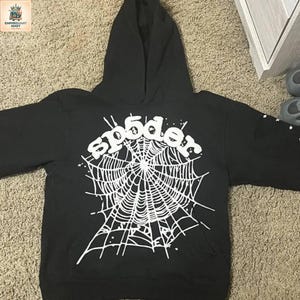 Puede incluir: Una sudadera con capucha negra que presenta un gran gr&aacute;fico blanco en la parte delantera. El dise&ntilde;o muestra un patr&oacute;n de telara&ntilde;a con el texto sp5der arqueado encima. Peque&ntilde;os detalles de estrellas blancas se esparcen por la red y la manga.
