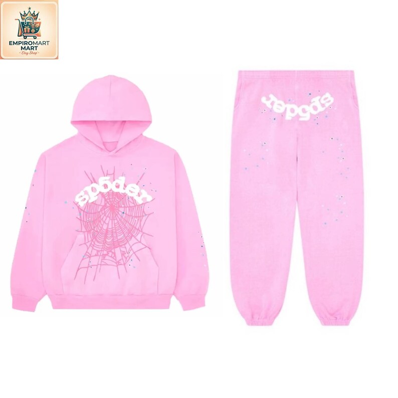 May include: A light pink hooded sweatshirt and matching sweatpants set. The hoodie features a white spiderweb graphic with the text "sp5der" in white. The sweatpants have the text "sp5der" in white on the upper back and small paint splatters.