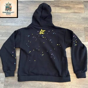 May include: A black hooded sweatshirt featuring a large yellow star graphic with the number 5 in the center on the upper back. The back and sleeves are decorated with a scattered pattern of small yellow, blue, and purple stars across the fabric.