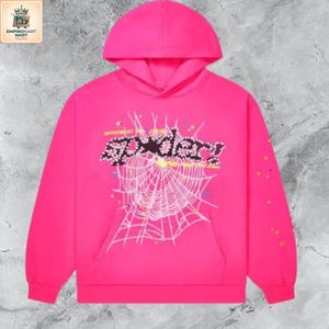 May include: A bright pink hooded sweatshirt featuring a large white spiderweb graphic on the front. The word spider is printed in a black, studded-style font across the web, accompanied by small colorful star accents and scattered dots.