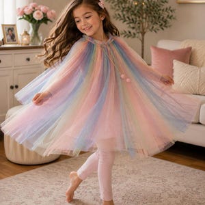 Könnte beinhalten: Ein regenbogenfarbener Tüllumhang mit gerafftem Halsausschnitt und zwei rosa Bommel-Bindebändern, getragen über einer hellrosa Leggings. Der transparente, mehrlagige Stoff zeigt einen Verlauf aus Pastelltönen mit dezenten Glitzerakzenten.