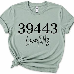 May include: A sage green short-sleeved t-shirt featuring the black printed text 39443 above a horizontal line, with the text Laurel, MS written in a cursive script font below the line. The shirt has a crew neckline and a knotted hem detail.