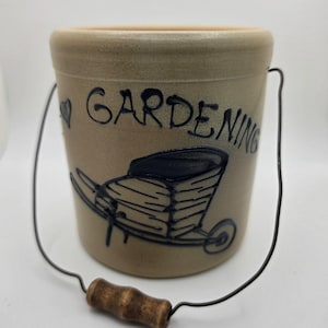 May include: A tan ceramic crock with a wire handle and a wooden grip. The front features a dark blue illustration of a wheelbarrow and the text "GARDENING" with a small heart symbol. The vessel has a cylindrical shape and a smooth finish.