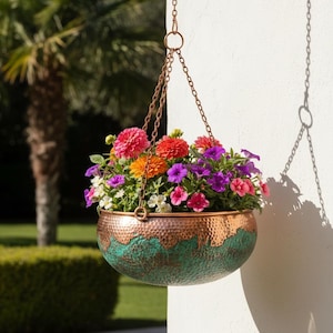 Op de afbeelding: Een ronde, gehamerde metalen hangende plantenbak met een koperkleurige bovenrand en een textuurrijke, verdigris-groene patina-afwerking op het onderste gedeelte. De bak hangt aan drie koperkleurige kettingen en is gevuld met felroze, oranje en paarse bloemen.