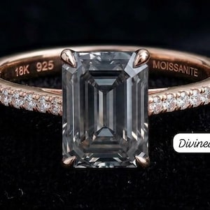 Op de afbeelding: Een ring met een grijze moissaniet in smaragdgeslepen vorm op een rosékleurige band. De band is aan de zijkanten bezet met kleine pavé-stenen en is aan de binnenzijde gegraveerd met de tekst 18K 925 en MOISSANITE. De ring is gelabeld Divinediamonds007.