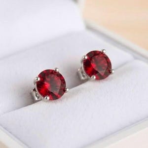 Può includere: Un paio di orecchini a lobo con pietre preziose rosso scuro a taglio rotondo, montate su griffe in metallo argentato. Ogni orecchino presenta una pietra circolare sfaccettata che riflette la luce, all'interno di un portagioie bianco.