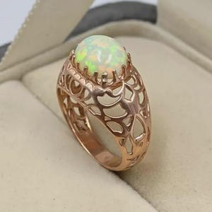 May include: A rose gold ring featuring an oval-shaped cabochon opal gemstone set in a prong setting. The band of the ring is crafted with an intricate, openwork filigree design that wraps around the finger, showcasing a detailed lattice pattern.