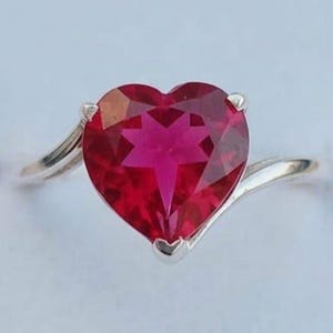 May include: A silver-toned ring featuring a large, heart-shaped, vibrant red gemstone with a faceted surface. The band has a smooth, polished finish that curves upward to meet the central stone. The text Top-Down Facet is visible below.