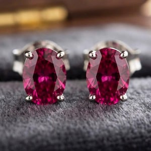 Könnte beinhalten: Ein Paar Ohrstecker mit oval geschliffenen, tief rosa-roten Edelsteinen in silberfarbenen Metallfassungen. Jeder facettierte Stein weist einen brillanten Glanz auf und ist auf einem dunkelgrauen, mit Samt ausgekleideten Untergrund platziert.