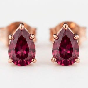 Puede incluir: Un par de pendientes de botón con gemas de color rojo intenso en forma de pera engastadas en metal de oro rosa. Cada gema está asegurada por tres garras, mostrando una superficie facetada, con los postes de metal visibles detrás de las piedras.