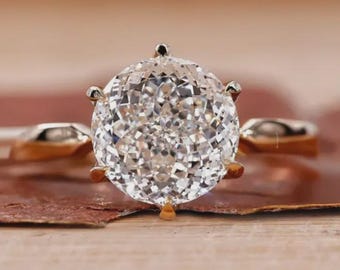 May include: A ring featuring a large, round, brilliant-cut clear gemstone set in a six-prong gold metal band. The stone displays numerous facets that reflect light, creating a sparkling appearance. The band is smooth and polished in a warm gold tone.
