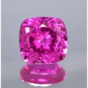 May include: A vibrant, cushion-cut gemstone in a deep, saturated magenta pink color. The stone features a complex, symmetrical faceted pattern that creates a brilliant, sparkling effect across its surface, reflecting light with high clarity.