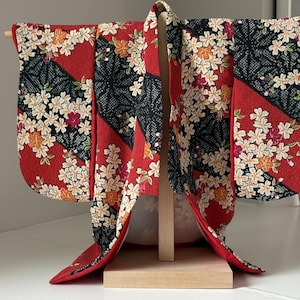 May include: A miniature kimono displayed on a wooden stand, featuring a vibrant red and black fabric with a repeating pattern of white cherry blossoms and small orange accents. The garment has wide sleeves and a structured collar design.
