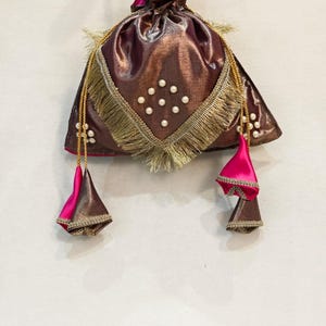 Puede incluir: Un bolso de mano tipo saco en bronce metalizado con una solapa triangular decorada con flecos dorados y perlas blancas. Incluye un asa de cuentas de perlas y dos borlas colgantes de tela rosa fucsia con bordes en bronce metalizado.