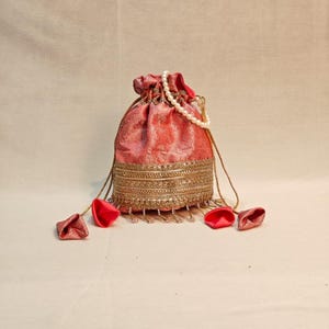 Op de afbeelding: Een koraalkleurig trekkoordtasje met een subtiel ton-sur-ton brokaatpatroon. De tas is versierd met een brede band van gedetailleerd goudkleurig borduurwerk, een handvat van witte parels en vier koraalkleurige stoffen kwastjes aan de koorden.