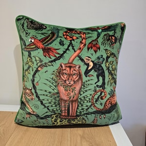 May include: A square throw pillow with a light green background featuring a detailed jungle print. The design includes a central orange feline, a red parrot, a monkey, and various tropical plants and vines, finished with green piping.