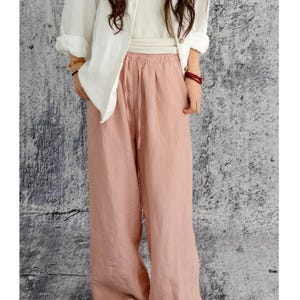 May include: Wide-leg trousers in a soft dusty pink colour featuring an elasticated waistband with a matching drawstring tie, side pockets, and a loose, flowing silhouette paired with a white long-sleeved button-down shirt and white open-toe sandals.