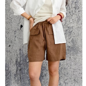 May include: Brown shorts featuring an elastic waistband with a drawstring, two large front patch pockets with flaps, and a relaxed fit. The shorts are paired with a white button-down shirt worn over a cream-coloured tank top.