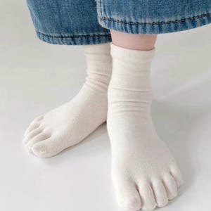 May include: A pair of off-white, ankle-length toe socks featuring individual compartments for each toe. The socks are made of a soft, ribbed knit fabric and are worn with blue denim jeans, extending slightly above the ankle bone for a snug fit.
