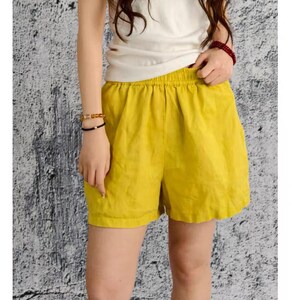 May include: Mustard yellow shorts featuring an elasticated waistband and a relaxed fit. The shorts are paired with a simple white sleeveless top. The fabric appears to have a textured finish, and the design includes a clean hemline at the bottom.