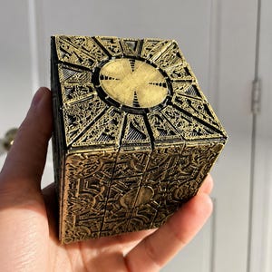 May include: A black and gold cube puzzle box featuring intricate, raised geometric patterns and engravings on every side. The surface displays a weathered, metallic finish with a central circular design element on each face of the cube.
