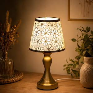 Peut inclure: Une lampe de table avec une base en métal doré lisse et un abat-jour en tissu blanc conique. L'abat-jour présente un motif répétitif de contours de fleurs dessinés à la main. La lampe est allumée, diffusant une lueur chaude depuis l'intérieur.