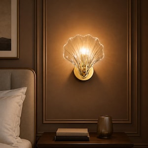 Peut inclure: Une applique murale dotée d'un abat-jour en verre transparent et nervuré en forme de coquillage. L'abat-jour est fixé à une base circulaire en métal doré qui se monte à plat contre le mur, diffusant une lumière chaude depuis l'intérieur.