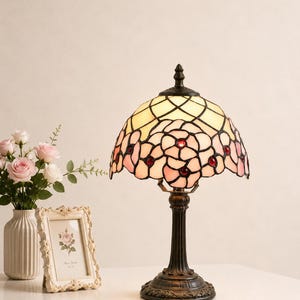 Peut inclure: Une lampe de table en vitrail avec un abat-jour en forme de dôme au motif floral crème et rose, agrémenté de petites perles de verre rouges. La lampe possède un pied en métal bronze foncé avec une colonne cannelée et un socle circulaire orné.